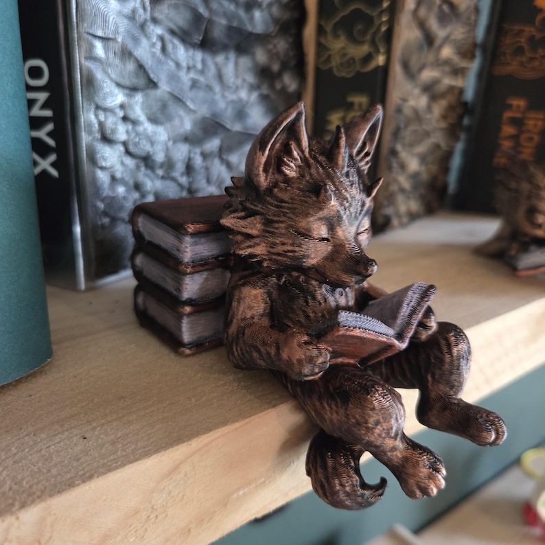 May include: A bronze-colored fox figurine reading a book, perched on a wooden shelf. The fox is sitting with its legs dangling, and a stack of miniature books is behind it. The word "ONYX" is visible on a book in the background.