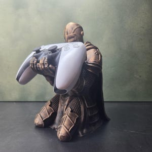 Puede incluir: Una figura de caballero de color bronce arrodillada que sostiene un mando de videojuego blanco y negro. El caballero lleva una armadura detallada con texturas de cota de malla y un casco, mientras que el mando presenta dos joysticks.