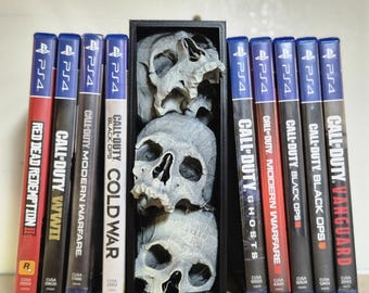 May include: A display of PS4 game cases, including "Red Dead Redemption II" and several "Call of Duty" titles. A black frame contains three grey skull sculptures. The games are arranged on a shelf.