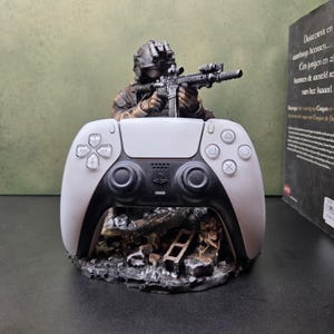 May include: A white and black wireless game controller resting on a detailed base featuring a soldier figure in tactical gear aiming a rifle. The base has a dark, weathered, rocky texture with debris, and the controller features standard buttons.