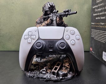 Könnte beinhalten: Ein weiß-schwarzer kabelloser Gamecontroller auf einem detaillierten Sockel mit einer Soldatenfigur in taktischer Ausrüstung, die mit einem Gewehr zielt. Die Basis hat eine dunkle, verwitterte Felsstruktur mit Trümmern und der Controller hat Tasten.