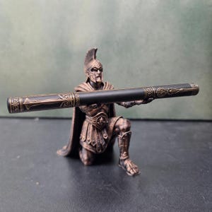 Puede incluir: Una figura de color bronce de un guerrero arrodillado con armadura y casco que sostiene un bolígrafo negro cilíndrico largo con intrincados patrones de filigrana dorados en los extremos. El guerrero está en posición de rodillas con los brazos extendidos.