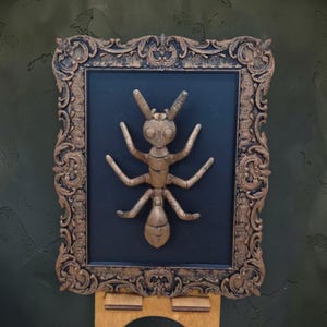 Op de afbeelding: Een gouden mierenbeeld in een sierlijke lijst op een zwarte achtergrond. De lijst heeft een gedetailleerd, vintage ontwerp. De mier is gecentreerd, met zes poten en antennes. Het kunstwerk wordt tentoongesteld op een houten ezel tegen een getextureerde, donkergroene muur.