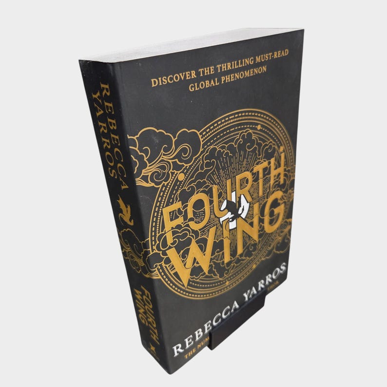 May include: A hardcover book titled "Fourth Wing" by Rebecca Yarros. The book has a black cover with gold lettering and design elements. The text "Discover the thrilling must-read global phenomenon" is also visible.
