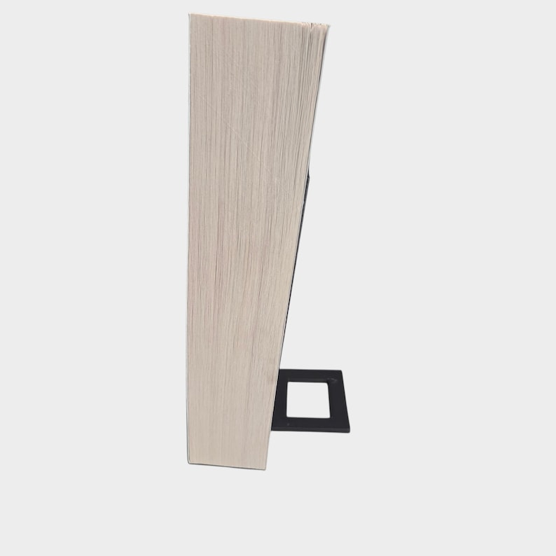 May include: A light-colored, rectangular wooden block, possibly a book, is propped up by a black, square-shaped stand. The wood grain is visible, and the object is set against a plain, light background.