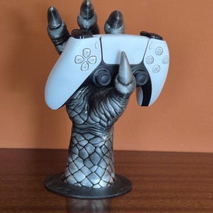 Op de afbeelding: Een grijze en zwarte drakenklauw-vormige controllerhouder die een witte PlayStation 5-controller ondersteunt. De klauw heeft een getextureerd, geschubd uiterlijk en een ronde basis. De controller is wit met zwarte accenten.