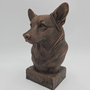 Può includere: Busto di cane color bronzo. La scultura presenta una dettagliata raffigurazione della testa e delle spalle di un cane, con orecchie prominenti e una superficie strutturata. Il busto è montato su una base quadrata.