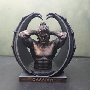 Peut inclure: Un buste aux tons bronze représentant une silhouette musclée aux cheveux attachés en chignon, encadrée par deux grandes ailes sombres et incurvées. La silhouette a les bras derrière la tête et repose sur une base rectangulaire marquée CASSIAN.