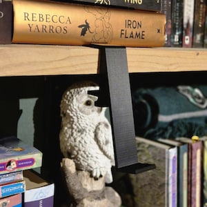 May include: A black metal bookend shaped like a bracket, holding up a book titled "Iron Flame" on a wooden bookshelf. A white owl figurine is in the foreground.