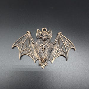 May include: A bronze-toned metal pendant shaped like a bat with outstretched wings. The bat features a detailed skull head, textured fur, and intricate scrollwork patterns on the wings. A small circular loop is attached to the top for hanging.