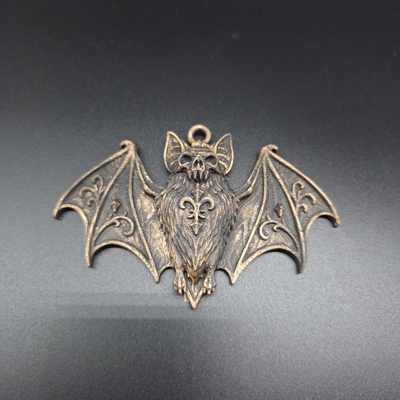 May include: A bronze-toned metal pendant shaped like a bat with outstretched wings. The bat features a detailed skull head, textured fur, and intricate scrollwork patterns on the wings. A small circular loop is attached to the top for hanging.