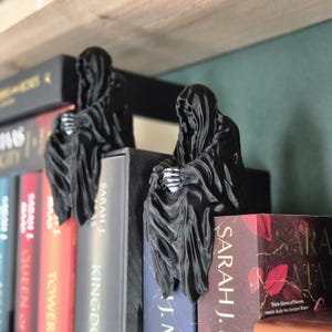 May include: Two black 3D printed bookends shaped like hooded figures, sitting on a bookshelf with a row of books. The bookends are holding books in place.