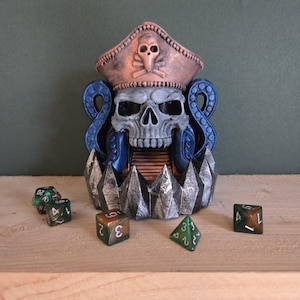 Può includere: Decorazione da tavolo a forma di teschio con cappello da pirata, tentacoli blu e base frastagliata. Diversi dadi verdi e marroni sono sparsi intorno alla base. Il teschio è grigio e il cappello è color bronzo.