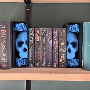 May include: A bookshelf arrangement featuring blue skull bookends with blue roses, flanking a row of books. Titles include "Twisted," "Scarred," "Wretched," "Hooked," "Crossed," and "Fourth Wing."