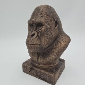 May include: A bronze-colored gorilla bust sculpture. The sculpture features a detailed depiction of a gorilla's head and upper chest, with a textured surface and a square base. The gorilla's facial features are clearly defined.