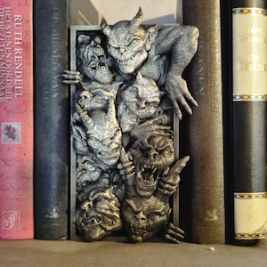 Op de afbeelding: Een 3D-geprint boekenlegger met een groep groteske, grijze en zilveren, demonische gezichten die uit een rechthoekig frame komen. De boekenlegger is geplaatst tussen twee boeken.