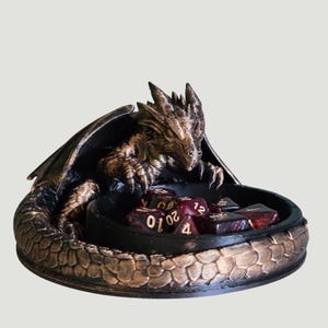 Peut inclure: Figurine de dragon en bronze tenant un bol noir rempli de dés. Le dragon a des écailles détaillées, des ailes et une expression féroce. Les dés sont rouge foncé avec des chiffres dorés, parfaits pour les jeux de société.