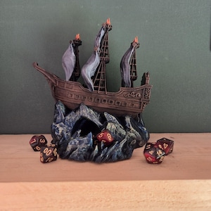 Può includere: Un modello di nave in miniatura marrone con tre alberi e vele grigie, caratterizzato da piccole fiamme arancioni sulla cima di ogni albero. La nave è montata su una base rocciosa blu scuro e nera, circondata da diversi dadi da gioco rossi e neri.