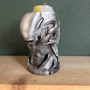 以下が含まれることがあります： エイリアンの頭の形をした銀色の缶クーラー。クーラーは、テクスチャ加工された表面、鋭い歯、リブのあるボディなど、詳細な機能を備えています。ソーダの缶が中に入っています。