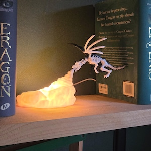 May include: A white skeletal dragon figurine with black accents, positioned to appear as if it is breathing a glowing, warm-toned light that emanates from a cloud-like base. The dragon is displayed between two books, one titled Eragon.