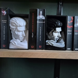 Könnte beinhalten: Ein Holzregal mit Büchern und dekorativen Büsten. Die schwarzen Bücher tragen weiße Titel wie "eclipse" und "morgenrood". Büsten männlicher Figuren stehen vor einigen Büchern. Eine ästhetische Präsentation.