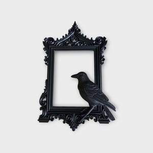 Peut inclure: Cadre photo noir orné avec un corbeau perché. Le cadre présente un design gothique détaillé avec une ouverture rectangulaire. Le corbeau est perché sur le bord du cadre, ajoutant une touche décorative. Le cadre et le corbeau sont noirs, créant un contraste saisissant.