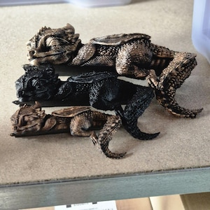 Op de afbeelding: Een verzameling gedetailleerde, miniatuur hagedisbeeldjes in verschillende kleuren. De beeldjes zijn in tinten bruin, zwart en brons, met een getextureerde huid en ingewikkelde details. Ze zijn gerangschikt op een neutraal oppervlak.