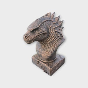 May include: A detailed bust of a dragon's head, in shades of brown and bronze. The sculpture features textured scales, a prominent snout, and sharp, spiky ridges along the head and neck. The dragon rests on a square base.