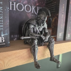 Könnte beinhalten: Eine silberfarbene Figur eines sitzenden Kriegers mit korinthischem Helm und Beinschienen, der ein Schwert auf dem Schoß hält. Die Figur ist in einer gebeugten, nachdenklichen Haltung dargestellt, mit detaillierter Rüstung und einem Umhang.
