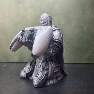 May include: A silver-toned knight figurine in armour kneeling while holding a white and black wireless video game controller. The knight features detailed chainmail textures and plate armour pieces, with the controller resting in its hands.