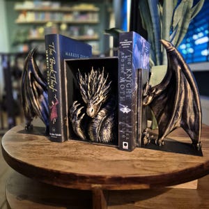 Könnte beinhalten: Buchstützen mit Drachenmotiv mit einer zentralen Drachenskulptur und zwei flügelförmigen Buchstützen. Der Drache ist gold und schwarz, und die Flügel sind bronze. Bücher werden zwischen den Buchstützen gehalten.