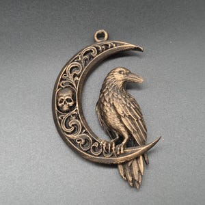 May include: A bronze-colored pendant featuring a crescent moon shape with intricate scrollwork and a small skull integrated into the design. A detailed raven is perched on the lower curve of the moon, which includes a small loop at the top.