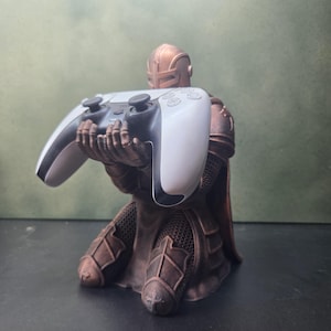 Pode incluir: Uma estatueta de cavaleiro cor de cobre ajoelhada segurando um controle de videogame sem fio branco e preto nas mãos. O cavaleiro usa uma armadura detalhada com capacete e textura de cota de malha, apoiando o controle com firmeza.