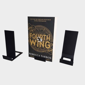 May include: A book titled "Fourth Wing" by Rebecca Yarros stands on a black metal book stand. Two additional black metal stands are on either side. The book cover is black with gold lettering and design.