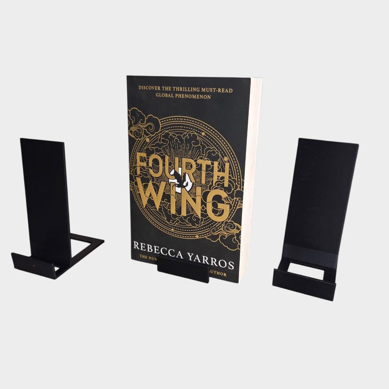 May include: A book titled "Fourth Wing" by Rebecca Yarros stands on a black metal book stand. Two additional black metal stands are on either side. The book cover is black with gold lettering and design.