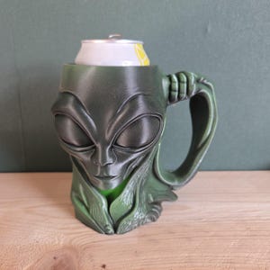 Può includere: Una tazza verde a tema alieno con manico, che contiene una lattina di soda. La tazza presenta un design dettagliato del volto alieno ed è realizzata in materiale verde scuro. La lattina è bianca con dettagli gialli.