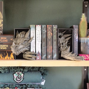 May include: A set of bookends featuring a dragon design. The bookends are made of a dark gray material and have a textured finish. The bookends are holding up a row of books with titles like "Twisted", "Scarred", and "Wretched".