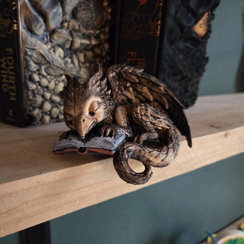May include: A detailed figurine of a sleeping griffin, with a book in front of it. The griffin is brown and tan, with detailed feather and scale textures. The book is open, with red-edged pages. The figurine is on a wooden shelf, with books in the background.