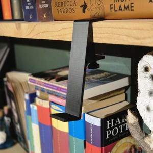 May include: A black metal bookend with a clamp design, holding up a stack of books on a wooden bookshelf. The bookend is attached to the shelf with a screw. The books on the shelf include titles like "Sky and Breath", "Blood", "Rebecca Yarros", and "Iron Flame".