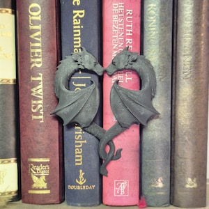 May include: A black metal dragon bookend in the shape of a heart. The dragons are facing each other and their tails are intertwined. The bookend is sitting on a bookshelf with other books.