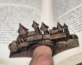Può includere: Un segnalibro a forma di castello color bronzo, con torrette dettagliate ed elementi architettonici. Il segnalibro è progettato per essere infilato su un dito, tenendo aperto un libro. Il design è intricato e sembra essere stampato in 3D.