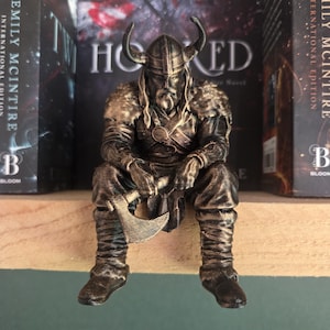 Op de afbeelding: Een bronskleurig beeldje van een zittende Vikingkrijger met een gehoornde helm en een schoudermantel met bonttextuur. Het figuur houdt een strijdbijl vast en is voorzien van gedetailleerde bepantsering, een gestructureerde broek en laarzen.