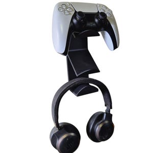 Op de afbeelding: Een zwarte wandbeugel voor een videogamecontroller en koptelefoon. De beugel heeft een witte PlayStation 5-controller eraan bevestigd. De koptelefoon is zwart met een zilveren accent.