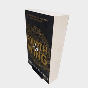 May include: A hardcover book titled "Fourth Wing" by Rebecca Yarros. The book has a black cover with gold lettering and a circular design. The text "Discover the Thrilling Must-Read Global Phenomenon" is at the top.