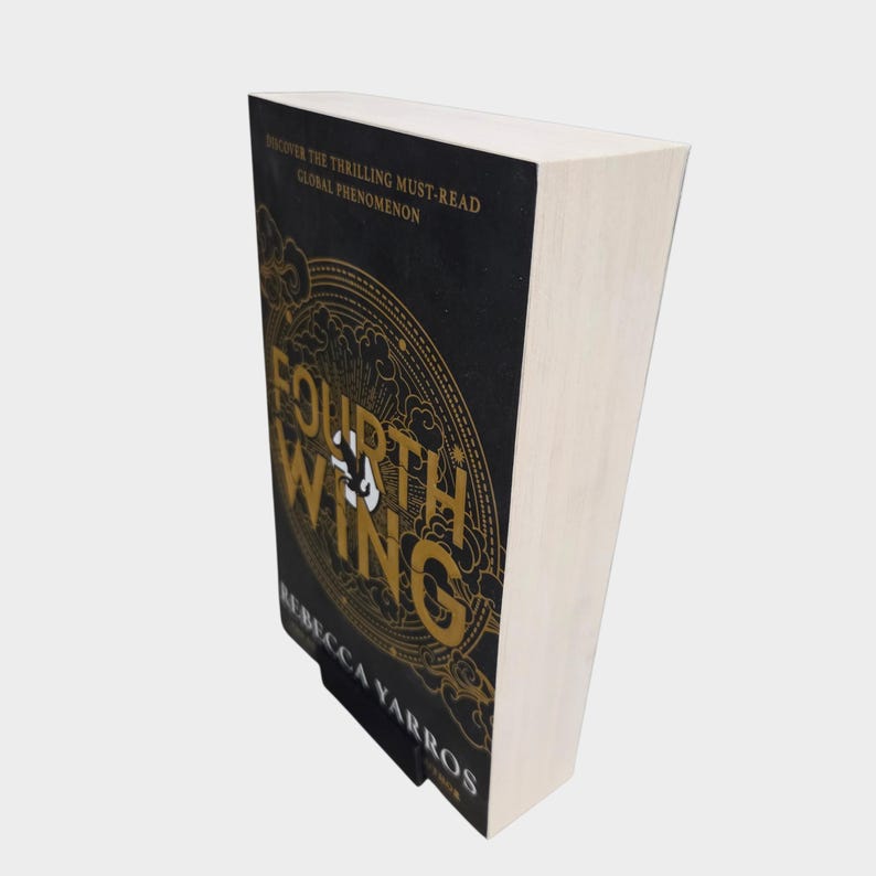 May include: A hardcover book titled "Fourth Wing" by Rebecca Yarros. The book has a black cover with gold lettering and a circular design. The text "Discover the Thrilling Must-Read Global Phenomenon" is at the top.