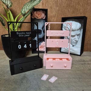 Op de afbeelding: Twee boektellers, één zwart en één roze, met de tekst "Books read this year". De zwarte teller toont "041" en heeft een lade. De roze teller toont "1" en heeft een lade. Twee kleine roze kaarten met cijfers liggen ervoor.