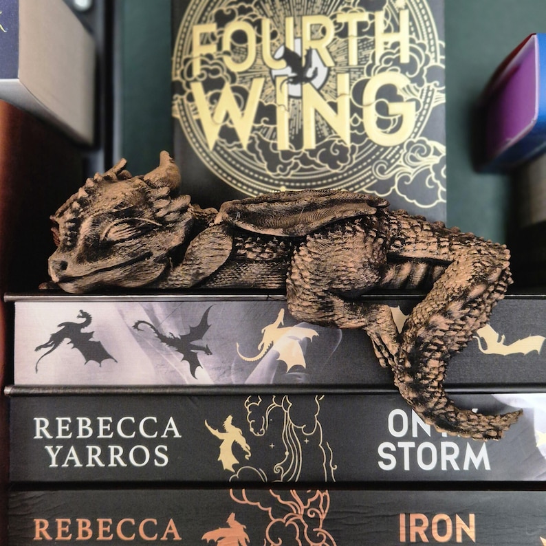 Op de afbeelding: Een bronzen drakenbeeldje rust op een stapel boeken. De boeken hebben titels als "Fourth Wing", "Rebecca Yarros" en "Iron Flame". De draak heeft gedetailleerde schubben en gesloten ogen, wat wijst op een ontspannen houding. Het thema is fantasie en literatuur.