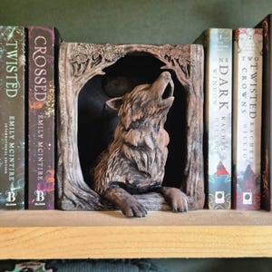 Op de afbeelding: Een gedetailleerde, bruine wolf-boekensteun, huilend in een met bomen omkaderde boog. De boekensteun staat op een houten plank, met verschillende boeken zichtbaar, waaronder "Twisted" en "Crossed" van Emily McIntire.