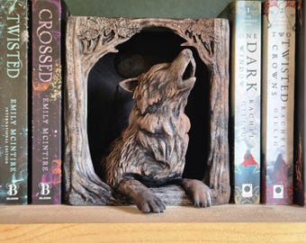 Puede incluir: Un sujetalibros detallado de un lobo marrón, aullando dentro de un arco enmarcado por árboles. El sujetalibros está colocado en una estantería de madera, con varios libros visibles, incluyendo "Twisted" y "Crossed" de Emily McIntire.