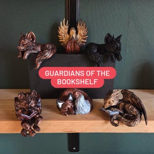 May include: A collection of fantasy bookshelf guardians. The figurines include a fox, a black cat, a griffin, and an angel with outstretched wings. The text "GUARDIANS OF THE BOOKSHELF" is displayed on a red banner.